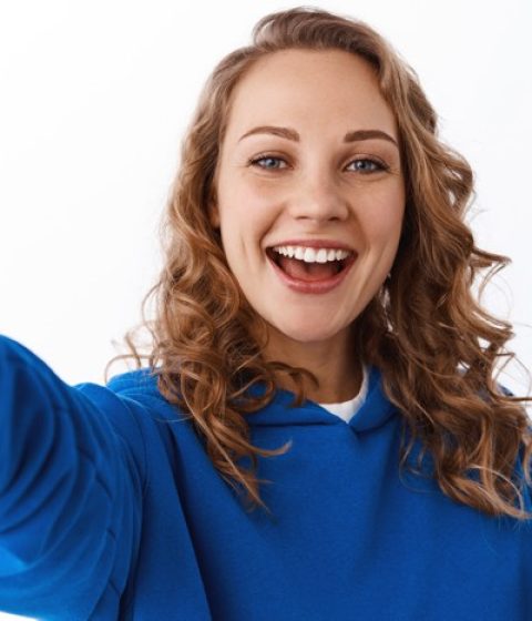Young happy beautiful girl taking selfie on mobile phone, stretch hand and hold smartphone, showing peace v-sign and smiling, white background.